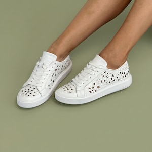 May include: White leather sneakers with intricate cut-out floral designs. The shoes have a low profile, white laces, and a thick white sole. The sneakers are shown on a light green background.