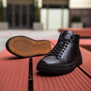 Puede incluir: Un par de zapatillas altas negras con parte superior de cuero liso, cordones negros y entresuela de goma negra. Una zapatilla está colocada en posición vertical, mientras que la otra está detrás, revelando una suela de goma color canela.