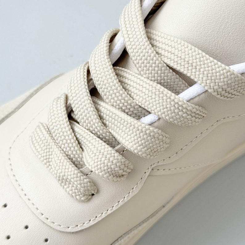 May include: A low-top sneaker in a solid cream color featuring a smooth leather upper with a perforated toe box. The shoe is secured with wide, flat, woven cream-colored laces that are threaded through eyelets in a classic crisscross pattern.