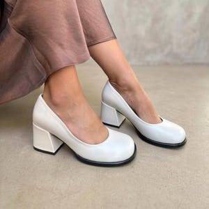 Op de afbeelding: Een paar off-white pumps met een ronde neus, een lage blokhak en een contrasterende zwarte zool. De schoenen hebben een gladde afwerking en een diepe uitsnijding, wat zorgt voor een strakke uitstraling bij een bruine rok.