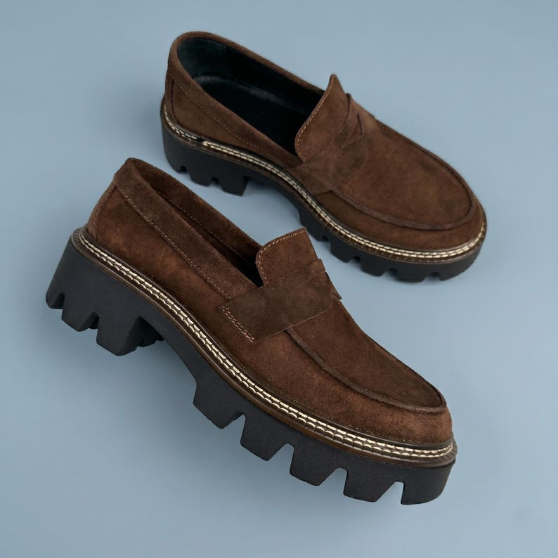 May include: A pair of brown suede loafers with a chunky black sole. The shoes have a classic penny loafer design with a stitched trim and a lug sole. The shoes are set against a light blue background.