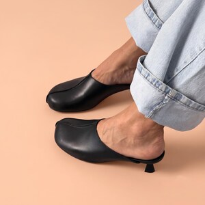 May include: A pair of black leather mules featuring a rounded toe, a central seam detail, and a small, low kitten heel. The footwear has an open back design and a smooth finish, paired with light blue denim jeans that are cuffed at the bottom.