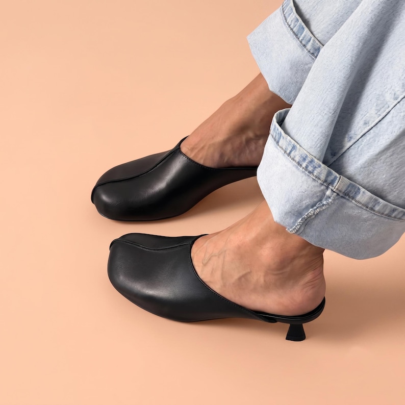 May include: A pair of black leather mules featuring a rounded toe, a central seam detail, and a small, low kitten heel. The footwear has an open back design and a smooth finish, paired with light blue denim jeans that are cuffed at the bottom.