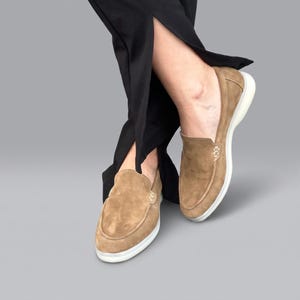 May include: A pair of tan suede loafers featuring a slip-on design with visible light-colored stitching along the vamp and a thick white rubber sole. The shoes are worn with black trousers that have a split hem detail at the bottom of the legs.