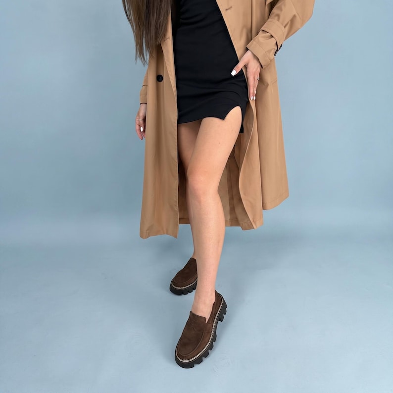 May include: A tan trench coat and a black dress are worn with brown loafers. The loafers have a dark brown suede upper, a thick black sole, and a light-colored stitch detail around the edge.