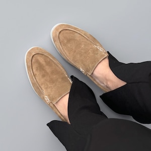 May include: A pair of tan suede slip-on shoes featuring a moccasin-style stitched toe seam and a clean white rubber sole. The shoes have a soft, textured finish and are worn with black trousers, showcasing a simple and functional footwear design.