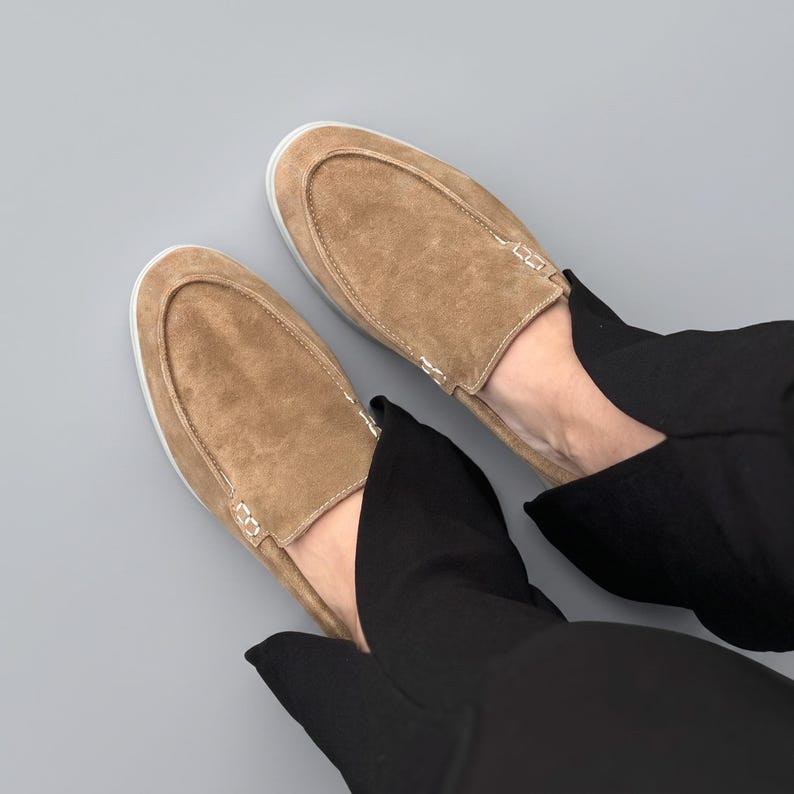 May include: A pair of tan suede slip-on shoes featuring a moccasin-style stitched toe seam and a clean white rubber sole. The shoes have a soft, textured finish and are worn with black trousers, showcasing a simple and functional footwear design.