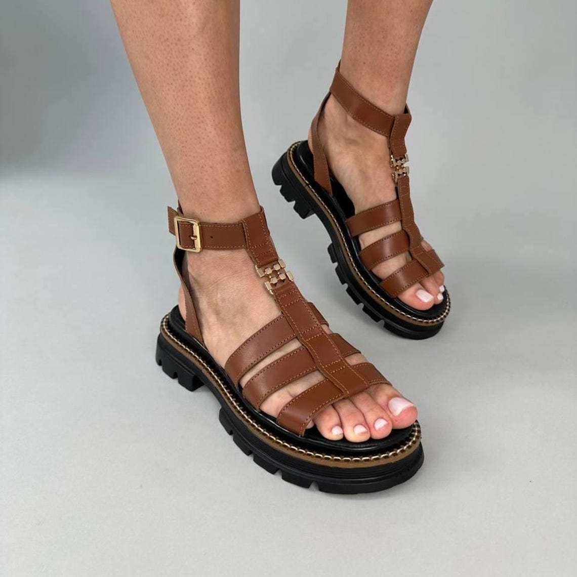 May include: Brown leather sandals with a T-strap design and a thick black sole. The sandals have gold-colored buckles and accents. The soles are approximately 1.5 inches thick.