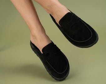 May include: Black suede slip-on loafers with a rounded toe and a low heel. The shoes have a classic design with a decorative strap across the top. The soles are black and the shoes are set against a green background.