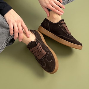 May include: A pair of dark brown suede sneakers featuring matching brown laces and a solid gum rubber sole. The low-top footwear has visible tonal stitching along the side panels and a rounded toe design, providing a clean and uniform appearance.