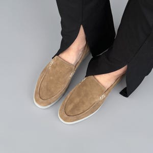 May include: A pair of tan suede slip-on loafers featuring a moccasin-style stitched toe seam and a white rubber sole. The shoes are worn with black trousers that have a small slit at the hem, showcasing the low-profile design of the footwear.