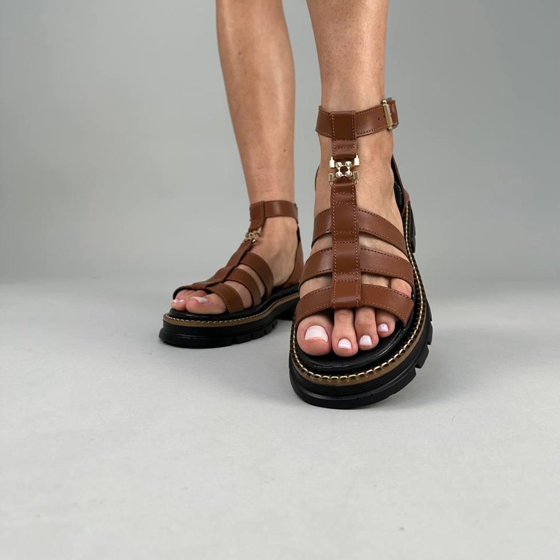 May include: Brown leather gladiator sandals with a chunky black sole. The sandals feature multiple straps and a buckle closure around the ankle. The soles have a height of approximately 1.5 inches.