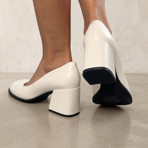 Op de afbeelding: Een paar off-white pumps met een ronde neus en een stevige, dikke blokhak. De schoenen hebben een gladde, effen buitenkant met een contrasterende zwarte zool die zichtbaar is aan de onderkant van de hak en de basis van de schoen.