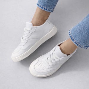 May include: A pair of white leather sneakers with white laces and a white sole. The sneakers have a classic design with perforations on the toe box. The shoes are worn with blue jeans.