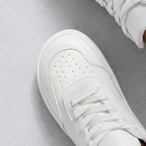 May include: A crisp white sneaker featuring a rounded toe box with a perforated ventilation pattern, tonal stitching along the side panels, and flat white laces threaded through the eyelets, showcasing a clean and uniform design.