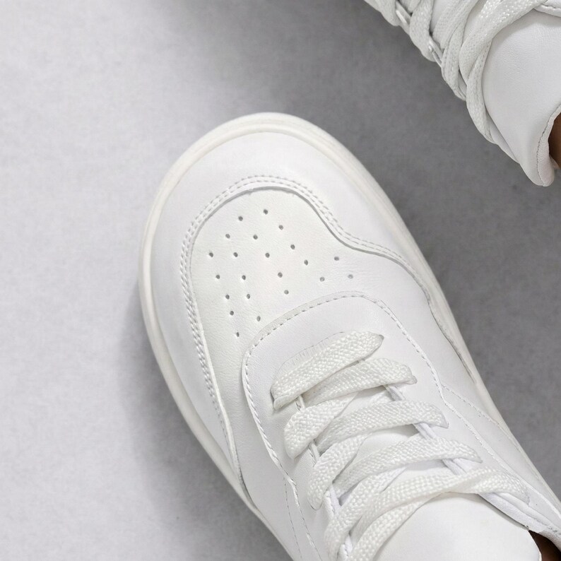 May include: A crisp white sneaker featuring a rounded toe box with a perforated ventilation pattern, tonal stitching along the side panels, and flat white laces threaded through the eyelets, showcasing a clean and uniform design.