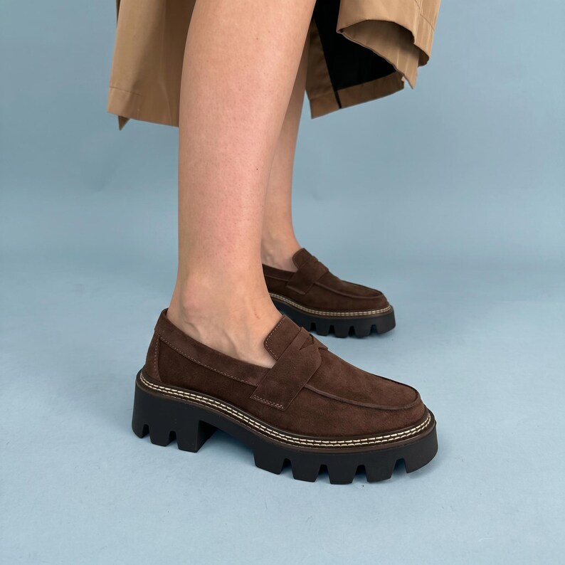 May include: Brown suede loafers with a chunky black sole. The shoes have a classic penny loafer design with a stitched trim. The soles are thick and have a rugged tread. The shoes are worn by a person.