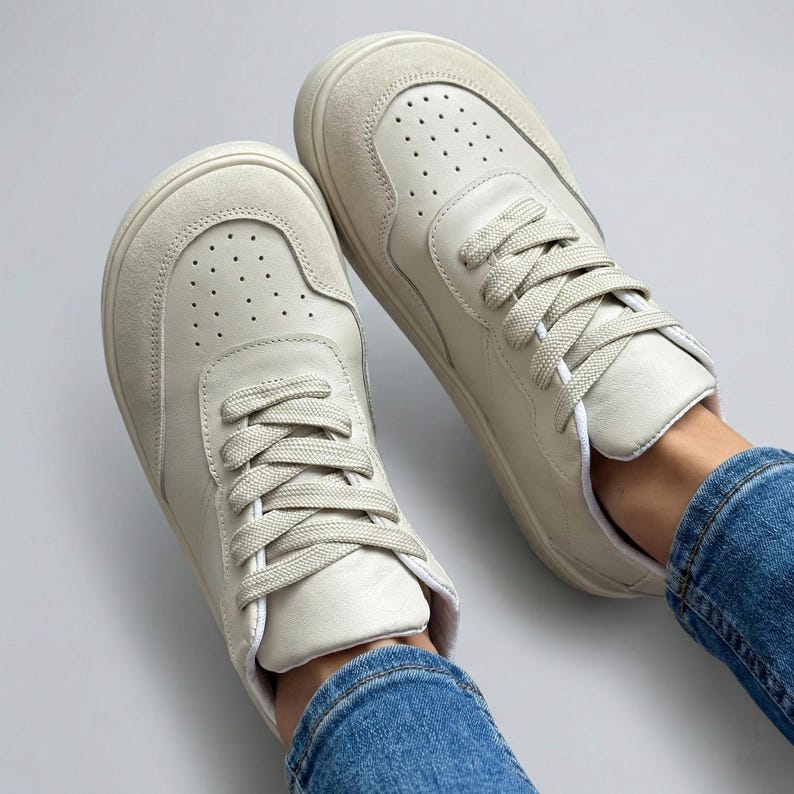 May include: A pair of off-white sneakers with matching laces. The shoes have a perforated toe box and a suede-like material on the upper. The sneakers are shown from a top-down perspective, with the laces neatly tied.