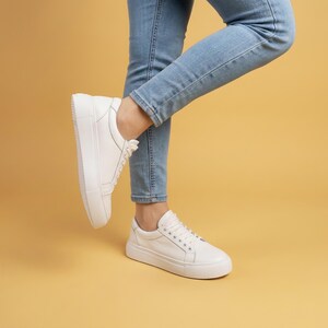 May include: White platform sneakers with laces. The shoes are worn with light blue jeans. The sneakers have a thick sole and a rounded toe. The background is a solid, warm yellow color.