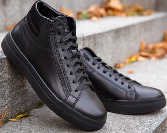 May include: A pair of black high-top trainers featuring a smooth leather-like finish, matching black laces, and a functional side zip closure. The shoes have a solid black rubber sole and a padded collar, providing a sleek and uniform appearance.