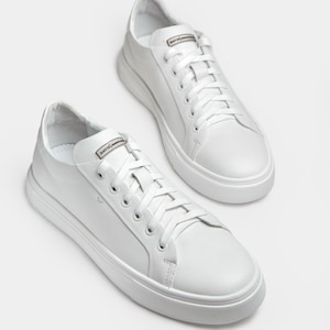 May include: A pair of white leather sneakers with white laces. The sneakers have a rounded toe and a thick white sole. The word "DAVIS" is printed on the tongue of each shoe. The shoes are displayed on a white background.
