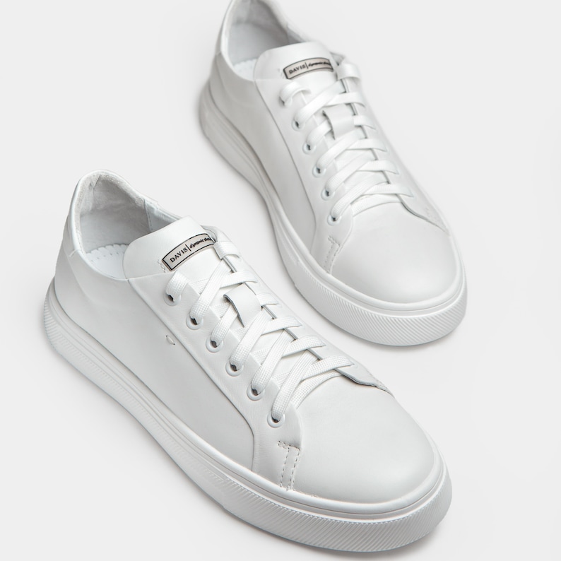 May include: A pair of white leather sneakers with white laces. The sneakers have a rounded toe and a thick white sole. The word "DAVIS" is printed on the tongue of each shoe. The shoes are displayed on a white background.