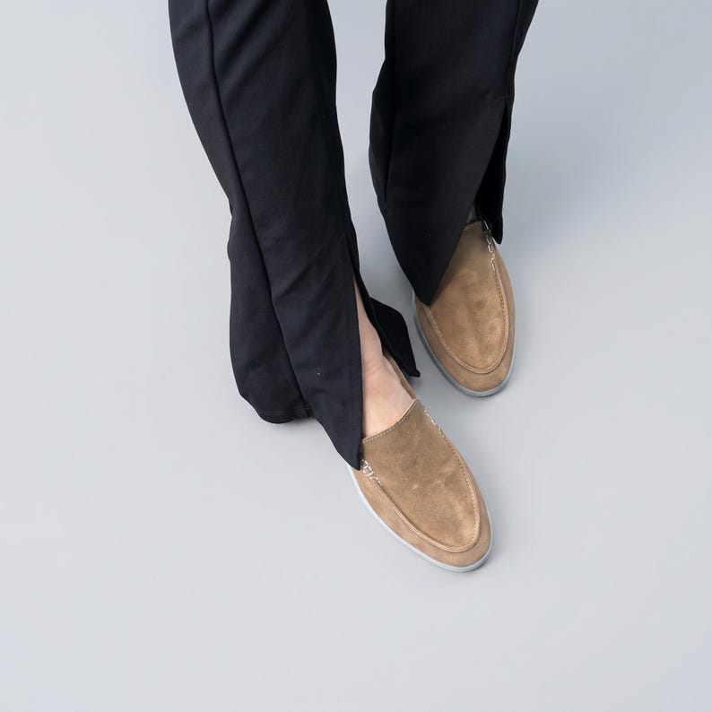 May include: A pair of tan suede loafers with visible white stitching along the moccasin-style toe seam, paired with black trousers that feature a vertical slit detail at the bottom hem, showcasing a clean and simple design aesthetic.