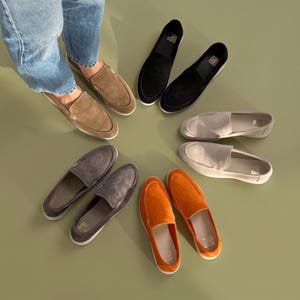May include: A collection of slip-on loafers arranged in a circle, featuring pairs in tan, black, light beige, orange, and dark grey suede. One pair of tan loafers is worn on feet with light blue denim jeans, while the others are displayed separately.