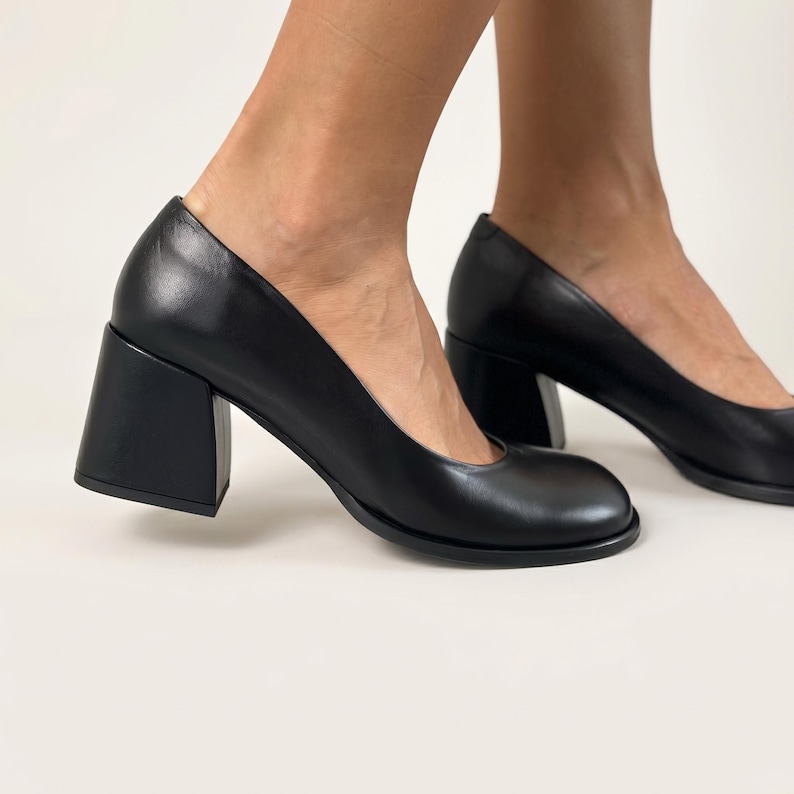 Op de afbeelding: Een paar zwarte leren pumps met een ronde neus en een dikke, robuuste blokhak. De schoenen hebben een gladde, matte afwerking en een laag uitgesneden bovenlijn, wat zorgt voor een strak en eenvoudig silhouet.