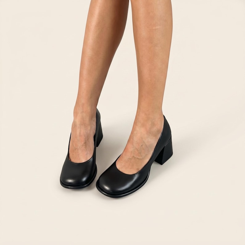 Op de afbeelding: Een paar zwarte leren pumps met een ronde neus, een gladde matte afwerking en een stevige blokhak. De schoenen hebben een diepe uitsnijding en een strak, onversierd ontwerp, wat zorgt voor een klassiek silhouet voor elke gelegenheid.