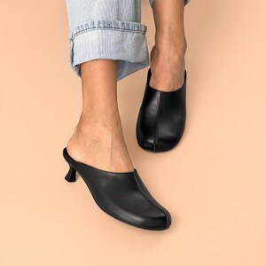 May include: A pair of black leather mules featuring a smooth finish, a rounded toe, a central seam detail running down the vamp, and a small, flared kitten heel. The footwear is designed as a slip-on style with an open back and a clean silhouette.