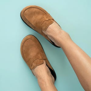 Op de afbeelding: Een paar beige suède instappers met een ronde neus, een gestikte voorzijde en een laag profiel. De schoenen hebben een gladde, matte textuur en een donkergekleurde binnenvoering, wat zorgt voor een eenvoudige en functionele schoenstijl.