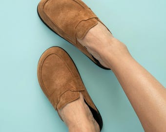 May include: A pair of tan suede slip-on loafers featuring a rounded toe box, a stitched apron front, and a low-profile design. The shoes have a smooth, matte texture and a dark-colored interior lining, providing a simple and functional footwear style.