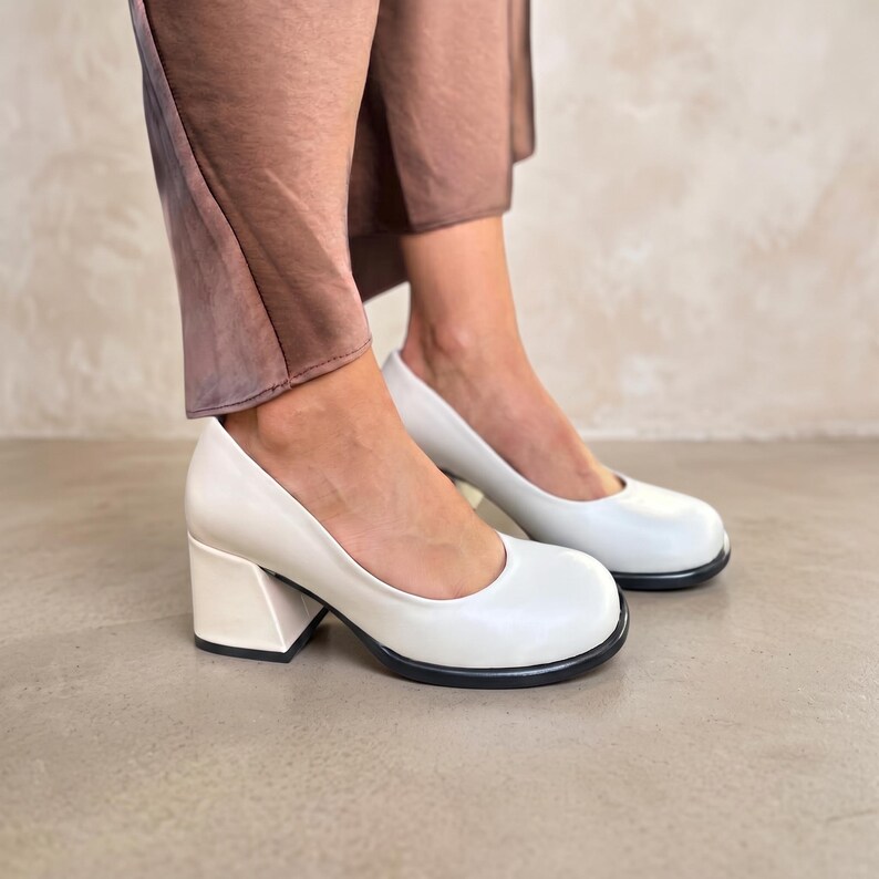 Op de afbeelding: Een paar off-white pumps met een ronde neus en een dikke blokhak. De schoenen hebben een glad, effen bovenwerk en een contrasterende zwarte zool, wat zorgt voor een strakke en eenvoudige uitstraling voor diverse gelegenheden.