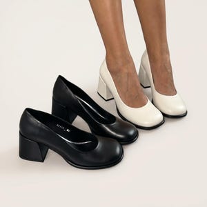 Op de afbeelding: Een paar zwarte pumps met blokhak en ronde neus en een paar off-white pumps met blokhak en ronde neus. De zwarte schoenen zijn voorzien van de tekst 60115 op de binnenvoering. Beide paren hebben een stevige, dikke hak.