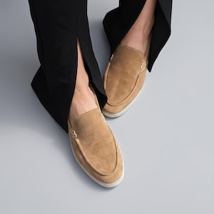 May include: A pair of tan suede slip-on loafers featuring a moccasin-style stitched toe seam and a white rubber sole. The shoes are worn with black trousers that have a side slit at the hem, showcasing the tan suede texture and clean design.