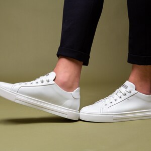 May include: White leather sneakers with a low-top design. The shoes have white laces and a white sole with a textured pattern. The sneakers are shown with black pants.