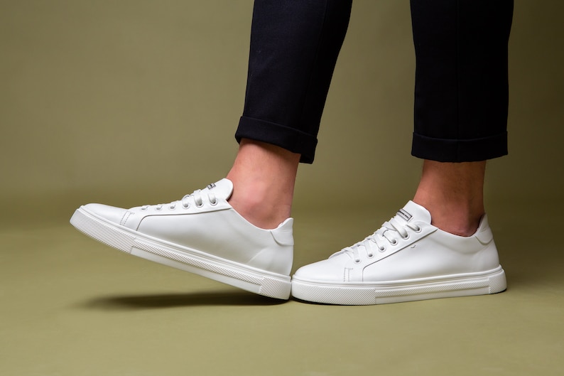 May include: White leather sneakers with a low-top design. The shoes have white laces and a white sole with a textured pattern. The sneakers are shown with black pants.