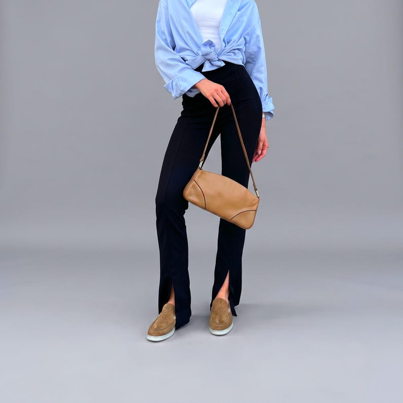 May include: A light blue button-down shirt tied at the waist, black high-waisted trousers with front hem slits, a tan leather shoulder bag with a thin strap, and tan suede loafers. The outfit is presented as a complete ensemble for daily wear.