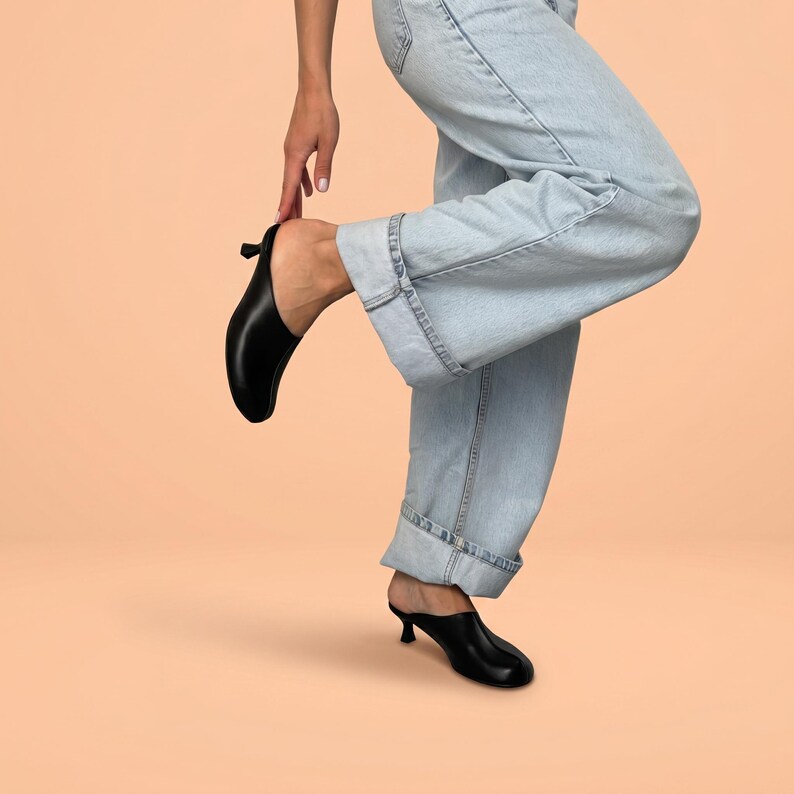 May include: A pair of black leather mules with a low kitten heel and a rounded toe, worn with light-wash blue denim jeans that feature a cuffed hem. The footwear has a smooth finish and a slip-on design, showcasing a clean and simple silhouette.