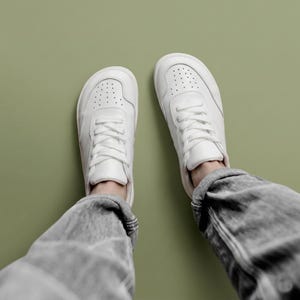 May include: A pair of white sneakers with matching white laces and a perforated toe box design. The shoes feature a rounded toe shape and are worn with grey denim jeans, showcasing a clean, low-top construction and smooth finish.