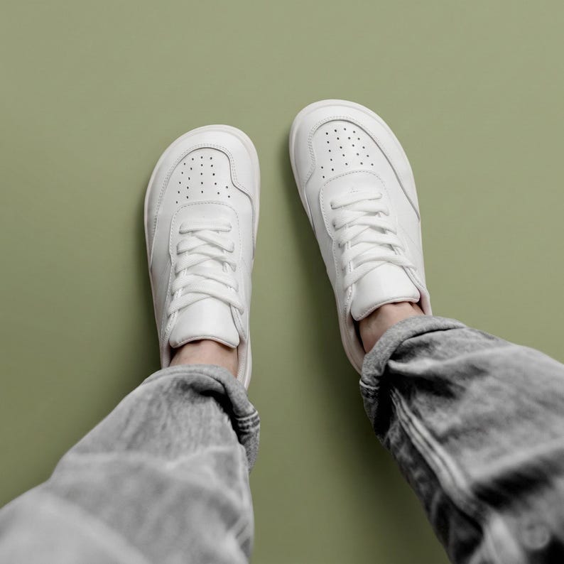 May include: A pair of white sneakers with matching white laces and a perforated toe box design. The shoes feature a rounded toe shape and are worn with grey denim jeans, showcasing a clean, low-top construction and smooth finish.