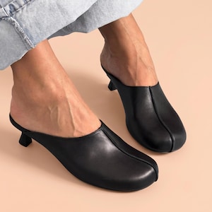 May include: A pair of black leather mules featuring a rounded toe, a center seam detail running along the top of the foot, and a low, flared kitten heel. The slip-on footwear has a smooth finish and a clean, structured silhouette for the feet.