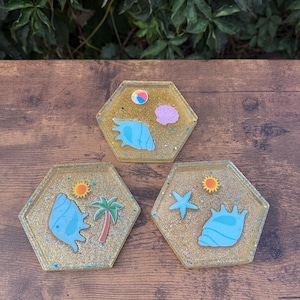 May include: Three hexagonal resin coasters with a gold glitter base. Each coaster features a different beach-themed design: a seashell, a beach ball, a starfish, a palm tree, and a sun. The coasters are arranged on a wooden surface.