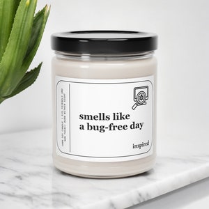 May include: A white soy candle with a black lid and a label that reads "smells like a bug-free day" with a computer screen and magnifying glass icon. The candle is on a white marble surface.