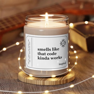 May include: A soy candle with brown wax and a white label that reads "smells like that code kinda works" with a small icon of a clock and a tick. The candle is on a wooden coaster and surrounded by white fairy lights.
