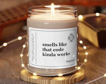 Op de afbeelding: Een sojawas kaars met bruine was en een wit label waarop staat "smells like that code kinda works" met een klein pictogram van een klok en een vinkje. De kaars staat op een houten onderzetter en is omringd door witte lichtsnoeren.
