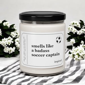 May include: A white soy candle with a black lid and a white label that reads "smells like a badass soccer captain inspired". The label also features a black and white soccer ball graphic.