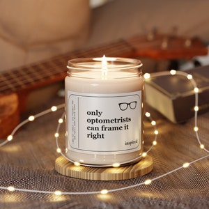 May include: A white candle with a glass jar and a label that reads "only optometrists can frame it right inspired". The candle is lit and sitting on a wooden coaster.