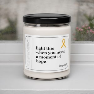 May include: A white soy candle with a black lid and a yellow ribbon. The label reads "light this when you need a moment of hope" and "inspired".